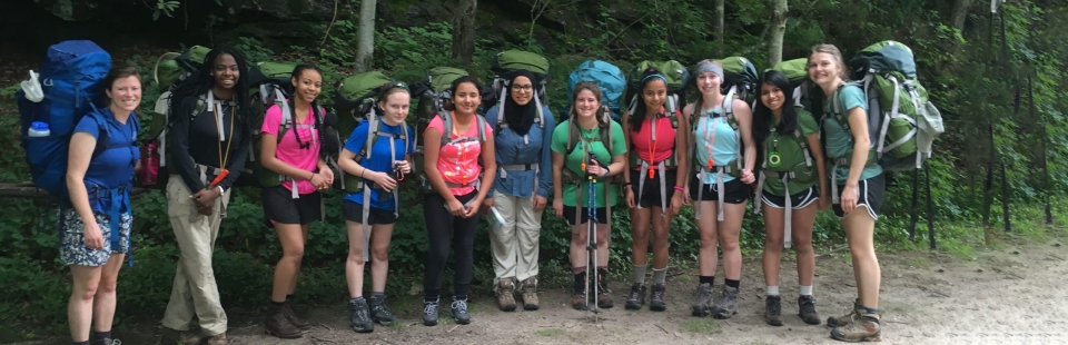 A group of participants in a past GALS program pose with their backpacks for a photo with a forest in the background.