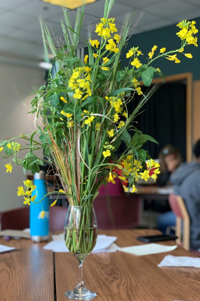 A vase filled with flowers including wheat and wood sorrel sit on a wooden table.