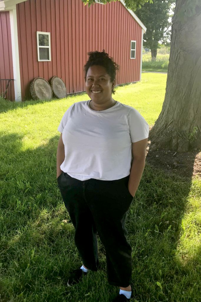 Rachel Richardson stands, smiling, in front of a tree on a summer day, wearing a white T-shirt and black pants.