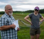Kevin Kahmark points to a diagram on a sheet of paper with another person looking on, while standing in a field at the W.K. Kellogg Biological Station.